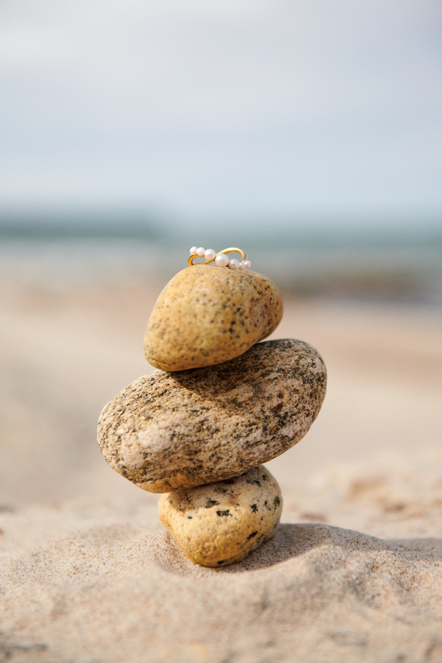 Pernille Corydon ring i forgyldt sølv med små perler, præsenteret på stablede strandsten i naturlige omgivelser, der symboliserer brandets inspiration fra naturen.