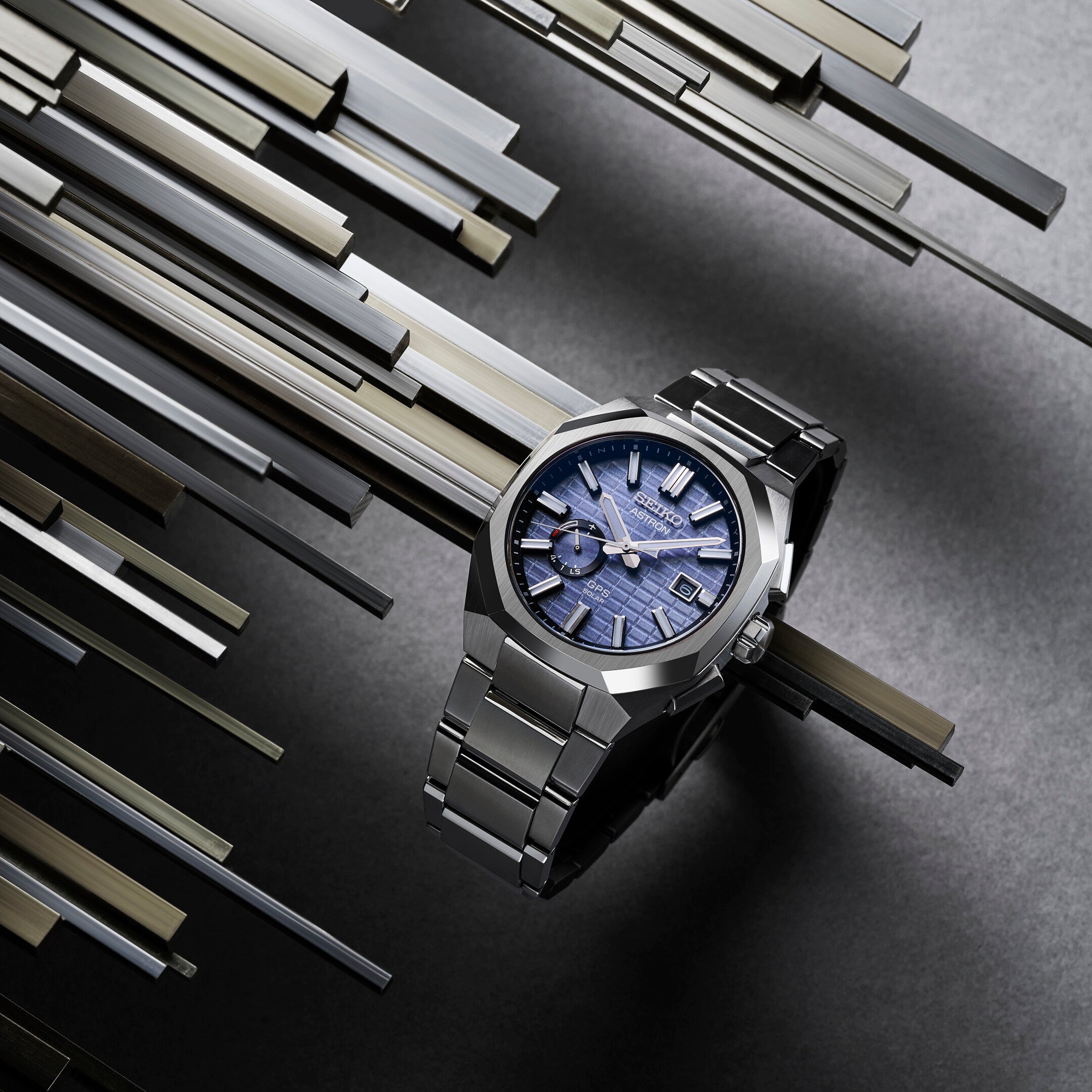Et Seiko Astron SSJ013 ur med blå urskive og rustfrit stålrem præsenteres på en baggrund af geometrisk arrangerede metalbjælker.