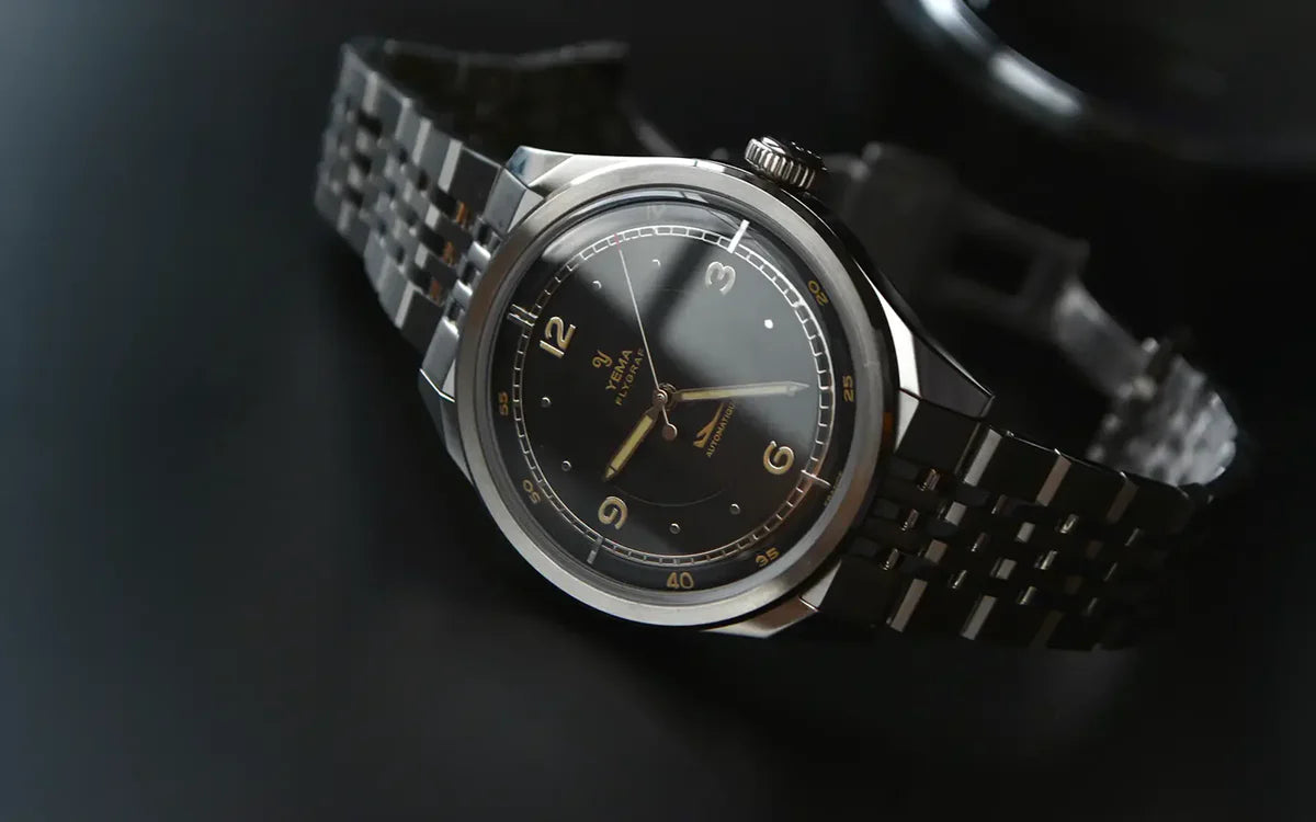 YEMA Flygraf Pilot Black armbåndsur i mørkt livsstilsbillede med sort skive, gyldne markeringer og stållænke.