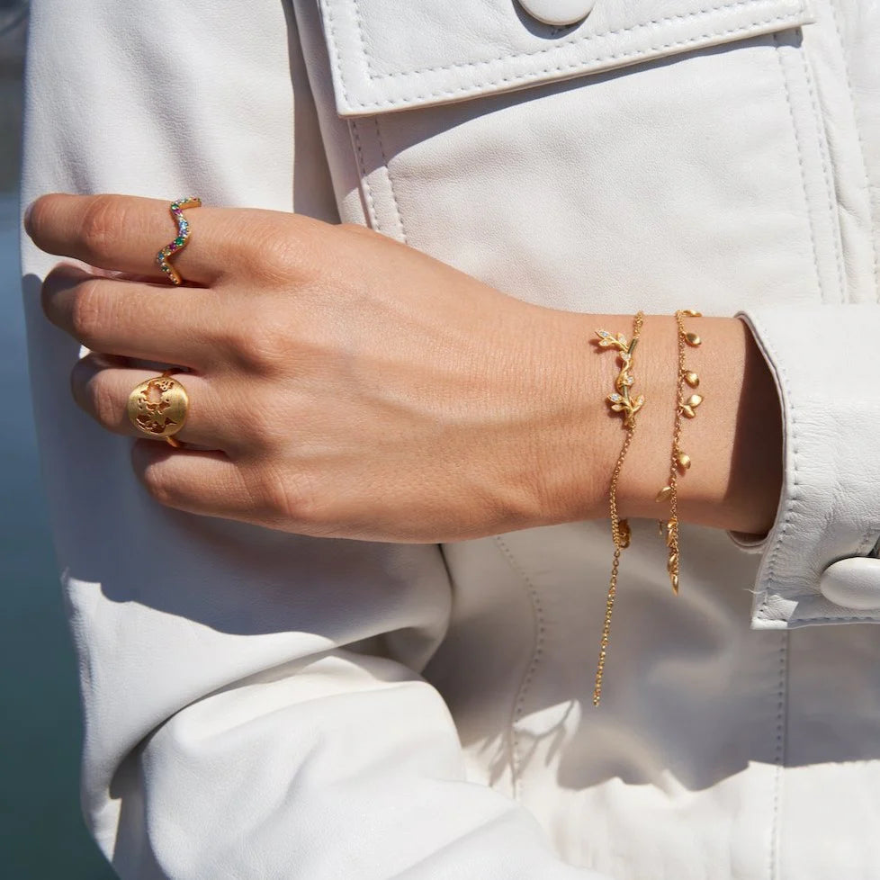  En person bærer to delikate guldbelagte armbånd fra ByBiehl, der elegant hviler på håndleddet. Det ene armbånd er udsmykket med fine blad- og grenmotiver, mens det andet har små, skinnende vedhæng. Begge armbånd udstråler feminin elegance og raffinement.