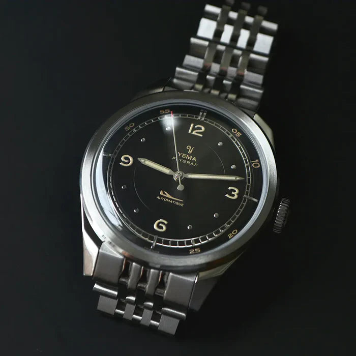 YEMA Flygraf Pilot Black armbåndsur i mørkt livsstilsbillede med sort skive, gyldne markeringer og stållænke.